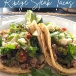 Tacos on a plate filled with ribeye steak, cilantro, cojita cheese and salsa. The plate is white and there are two tacos with two corn tortillas each.