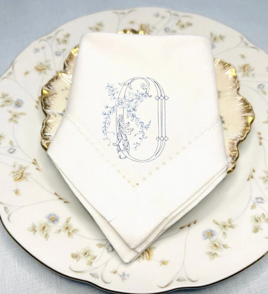 A beautiful blue and white spring tablescape embroidered dinner napkin in white stitched in two different blues set on top of a mismatched china place setting with soft colors and a gold rim on the top plate
