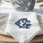 A table set with a white dinner napkin with embroidered "C" in blue sits on top of a natural fibre placemat with a white plate and chinoiserie vase.