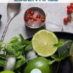 A top view of a bar mixing spoon and a jigger with berries, mint, and lime on a gray granite table.