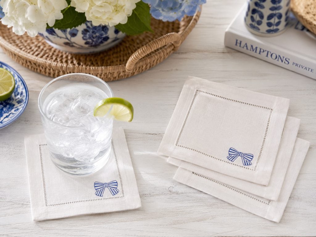 White linen embroidered cocktail napkins on a home bar next to a cocktail. The embroidered design is a blue striped bow.