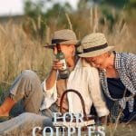 A couple sitting in a meadow sharing a bottle of wine. The couple is two women with matching hats in neutral colors laughing. Romantic picnic date ideas for everyone.