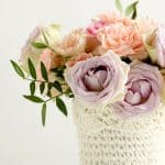 A bouquet of pastel colored roses and greens in a crocheted covered pot.