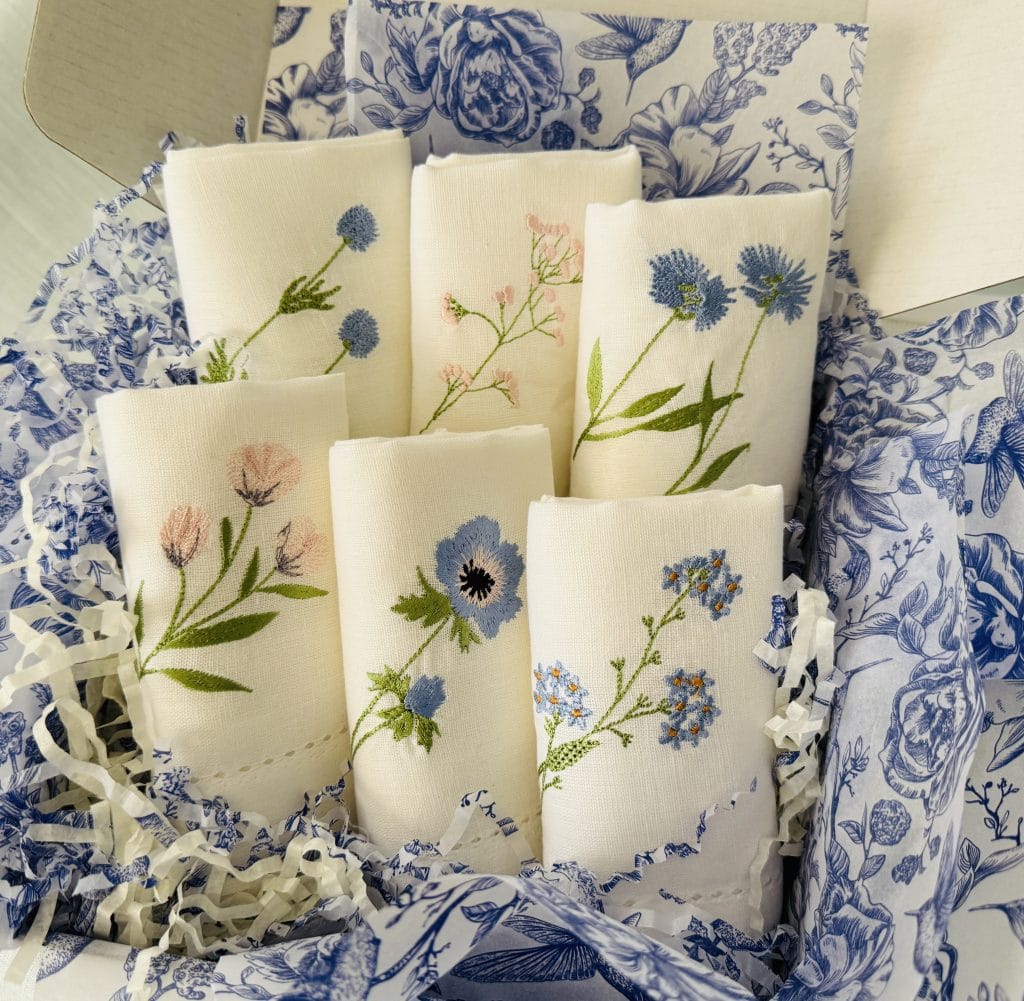 Set of 6 white dinner napkins embroidered with different wildflowers. Set in a gift box with blue and white tissue paper.