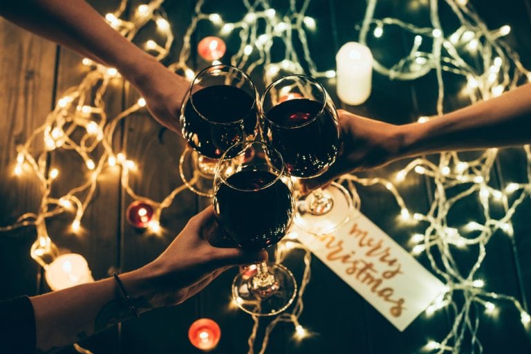 Three friends clinking glasses with red wine over some white lights with a sign that says Merry Christmas.