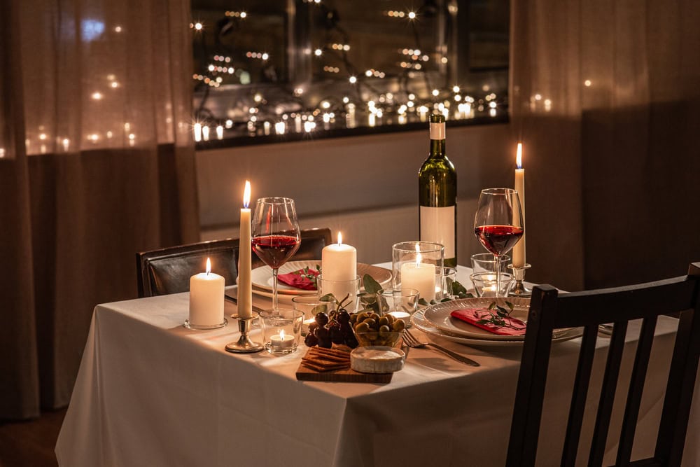 Close up of Valentine's Day dinner table serving for two with red wine in glasses, snacks and candles burning at home on valentine's day. The table is in front of a window looking out at the cityscape.