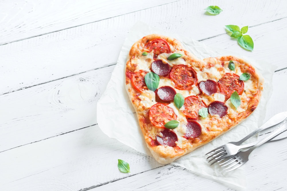 A heart shaped pepperoni and tomato pizza sitting on parchment paper on a white wooden table with forks.