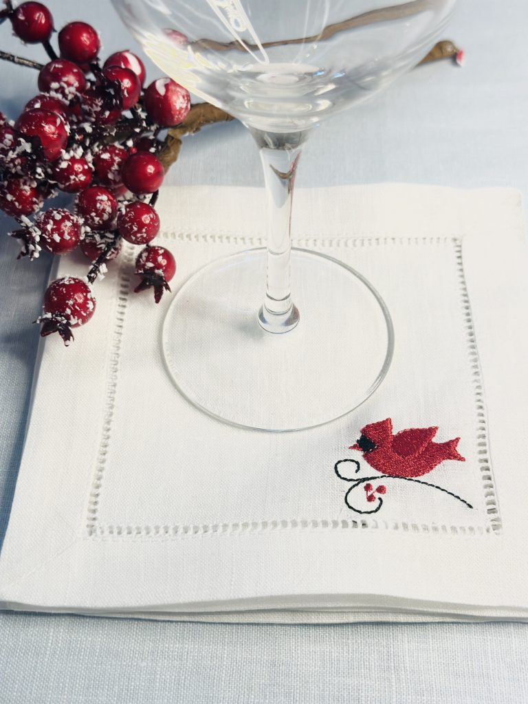 A simple red cardinal on a branch embroidered on the bottom right side inside the hemstitch. The napkin is bright white and there is a wine glass resting on it along with red berry branch next to them.