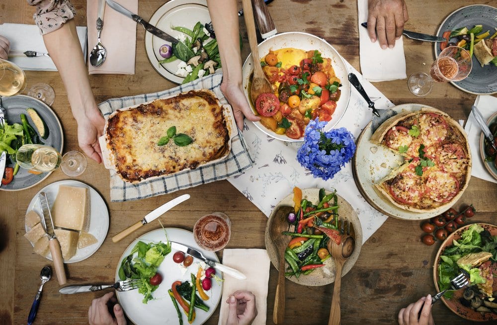 A dinner party table with lasagna, salads, cheese and other shared dishes. You see the people's hands who are at the table.