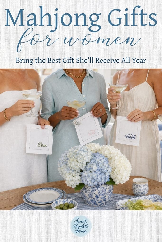 Three women standing next to a table at a party. You see from shoulders down. They are holding embroidered linen oversized coasters that have the words Bam, Dot, Crack embroidered on them.